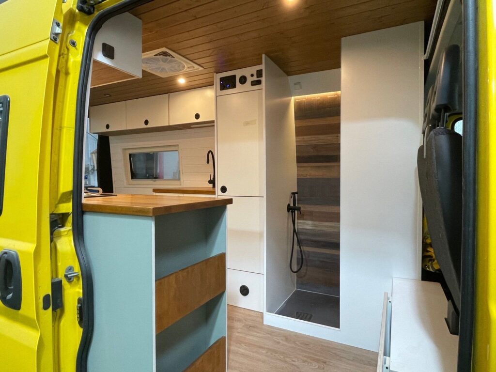 Interior de una Peugeot Boxer L3H2 2+2 camper, vista desde la puerta trasera, con mueble de almacenamiento y cocina a la izquierda y área para dormir a la derecha.