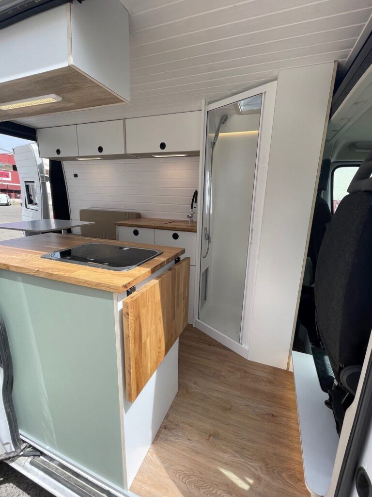 Interior moderno y funcional de una camper Fiat Ducato L2H2 diseñada por Baovan Camper para dos personas, destacando su cocina personalizada con encimera de madera y detalles en color verde pastel, armarios superiores y un espacio eficiente y elegante para vivir la aventura sobre ruedas con todas las comodidades del hogar