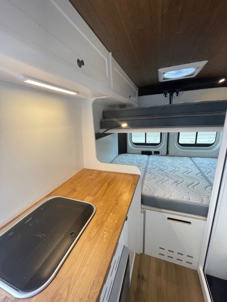 Detalle de la encimera con las camas al fondo, armarios blancos y encimera de madera en la furgoneta Camper Peugeot Boxer L3H2 4+4.