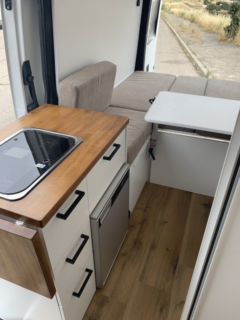 Acabado blanco y amadeirado de la cocina en la Camper Mercedes Sprinter L3H2 4+4. La foto muestra la zona del fregadero, con nevera, cajones y sofás detrás con una mesa extensible. Camperizado por BaovanCamper.