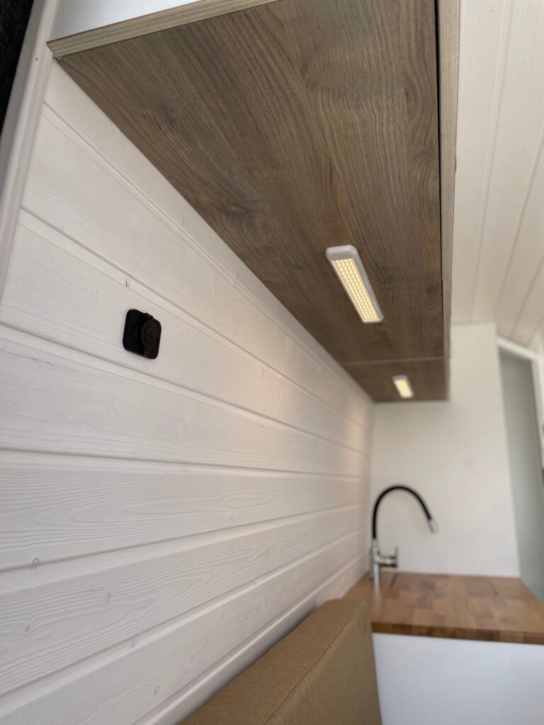 Detalles de la iluminación sobre el fregadero, de LED, con paredes blancas y acabado de madera en la Camper Fiat Ducato L2H2 para 2, convertida por Baovan Camper.