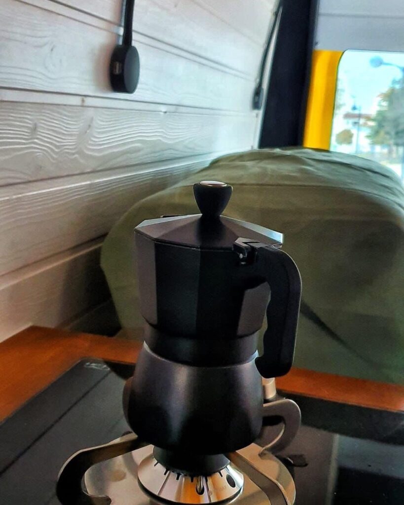 Vista de una cafetera italiana de color negro sobre una estufa de gas, con el interior de la furgoneta camperizada Boxer L3H2 2+2 de fondo, destacando el contraste entre la modernidad de los utensilios y el diseño rústico del vehículo.