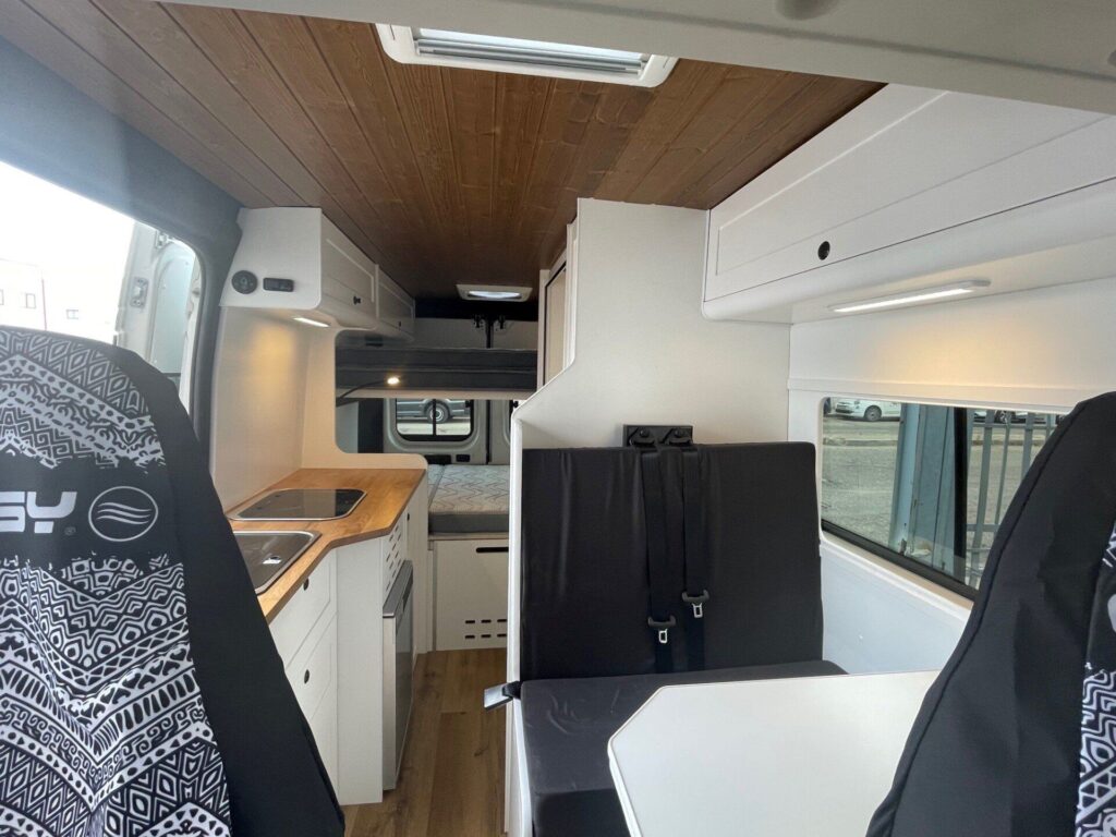 Vista desde el asiento del conductor de la Camper Peugeot Boxer L3H2 4+4, mostrando todo el interior de la furgoneta con espacio para cocina, fregadero, encimera, armarios, zona de descanso y cama al fondo. Amplio y con camas estilo litera (dos camas dobles).