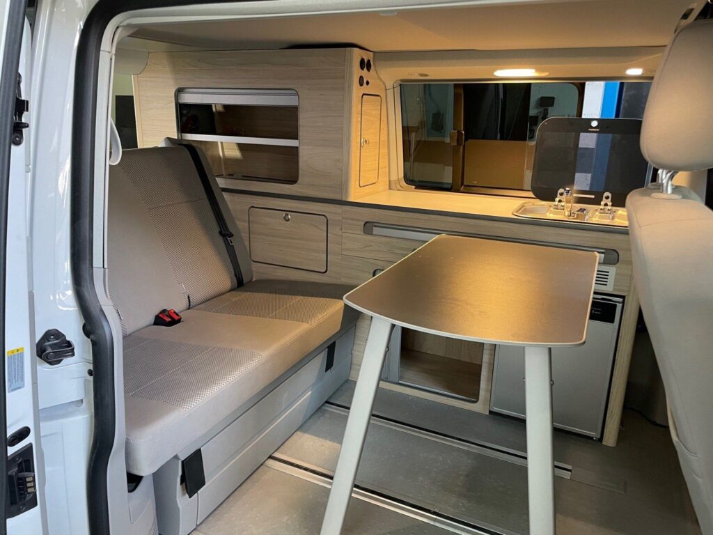 Espacio de asientos en la Camper California T6 convertido en área de comedor con mesa desplegable y asiento habilitado.