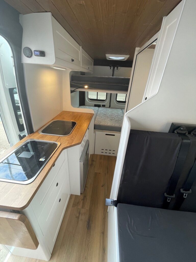 Vista aérea de la encimera con fregadero y cocina, asientos de la zona de descanso y camas al fondo en la furgoneta Camper Peugeot Boxer L3H2 4+4.
