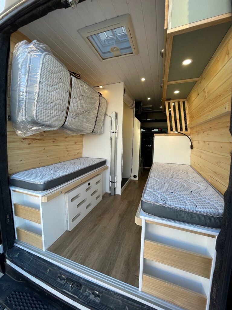 Detalle de las camas cerradas. Hay dos camas (o asientos) en las laterales y colchones para cerrar la cama de casal colgados en la pared, acabado blanco y de madera en la Iveco Daily 4+4 Pawlonia. Camperizado por BaovanCamper