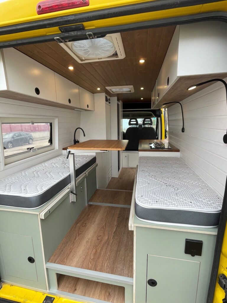 Vista del área de comedor convertible en cama dentro de una Peugeot Boxer L3H2 2+2, con asientos grises y mesa plegable.
