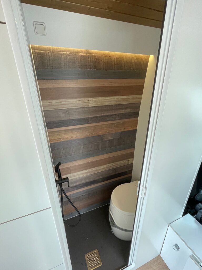 Vista interior del baño en una furgoneta camperizada Boxer L3H2 2+2. Se observa un inodoro de color crema a la derecha y una ducha con grifería de color negro a la izquierda. La pared del fondo está revestida con paneles de madera en tonos marrones. El piso es de color gris oscuro y hay una rejilla de drenaje dorada. La puerta del baño está abierta mostrando parte del exterior del baño que tiene azulejos blancos.