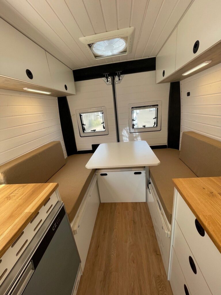 Interior de una camper Fiat Ducato L2H2. Se observa una área de estar con dos bancos opuestos y una mesa blanca retráctil en el centro, lo que proporciona un espacio práctico para comer o trabajar. Encima de la mesa, hay una claraboya que permite la entrada de luz natural y ventilación. La iluminación adicional viene dada por lámparas instaladas debajo de los armarios superiores, que aportan un toque moderno y funcional.