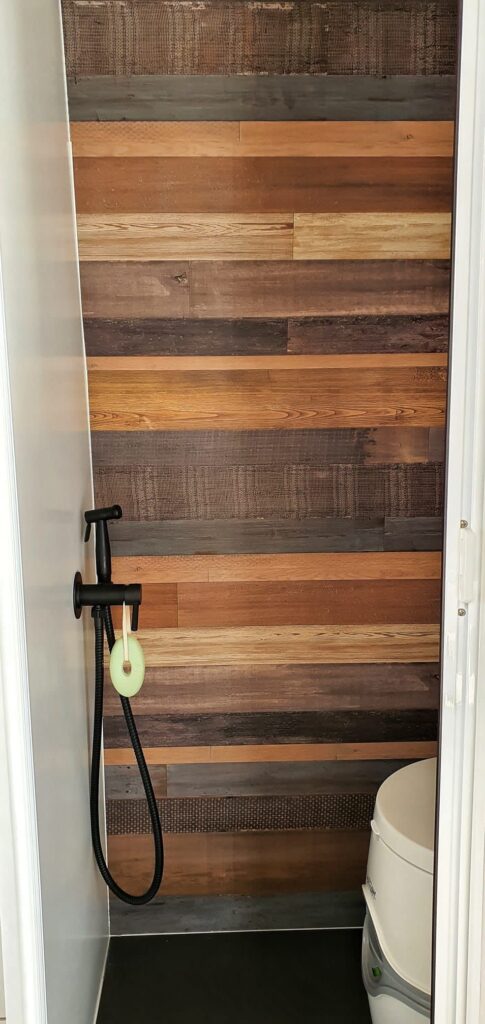 Detalle del baño en la furgoneta camperizada Boxer L3H2 2+2, con una ducha de mano de color negro montada en una pared de madera de diferentes tonalidades y un inodoro blanco en la esquina.
