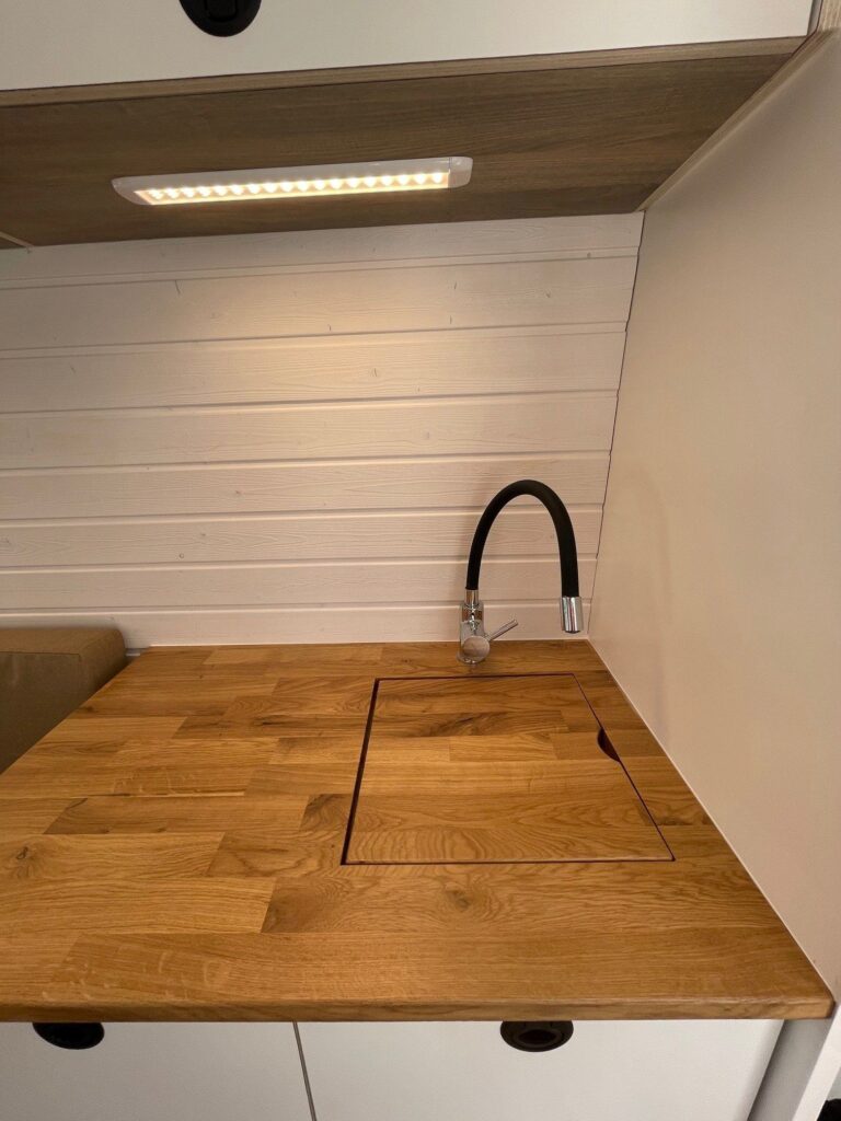 Detalle del fregadero, con una tapa que aumenta el espacio de trabajo, en la Camper Fiat Ducato L2H2 para 2, convertida por Baovan Camper. Pared blanca con acabado elegante.