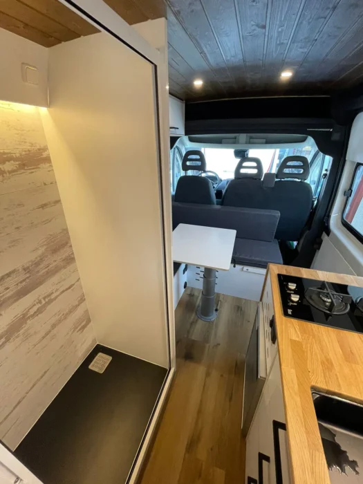 Interior de Camper L3H2 mostrando la vista desde la cocina hacia la cabina con una configuración de salón en 'L' para dos personas y suelo de madera.
