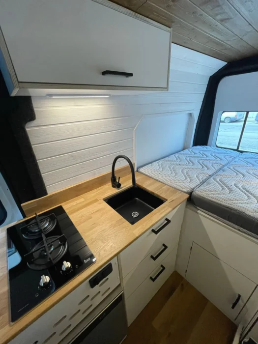 Cocina compacta en una camper L3H2 con fregadero de acero inoxidable, encimeras de madera y cocina de gas, bajo iluminación LED suave.