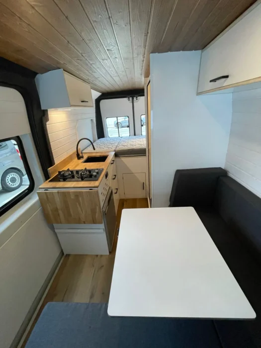 Área de comedor en una camper L3H2 con asientos en 'L', mesa amplia y armarios superiores, mostrando un ambiente acogedor y funcional.