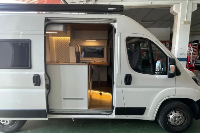 Vista exterior de una Peugeot Boxer Camper con puertas correderas abiertas, mostrando un interior acogedor iluminado y listo para la aventura.