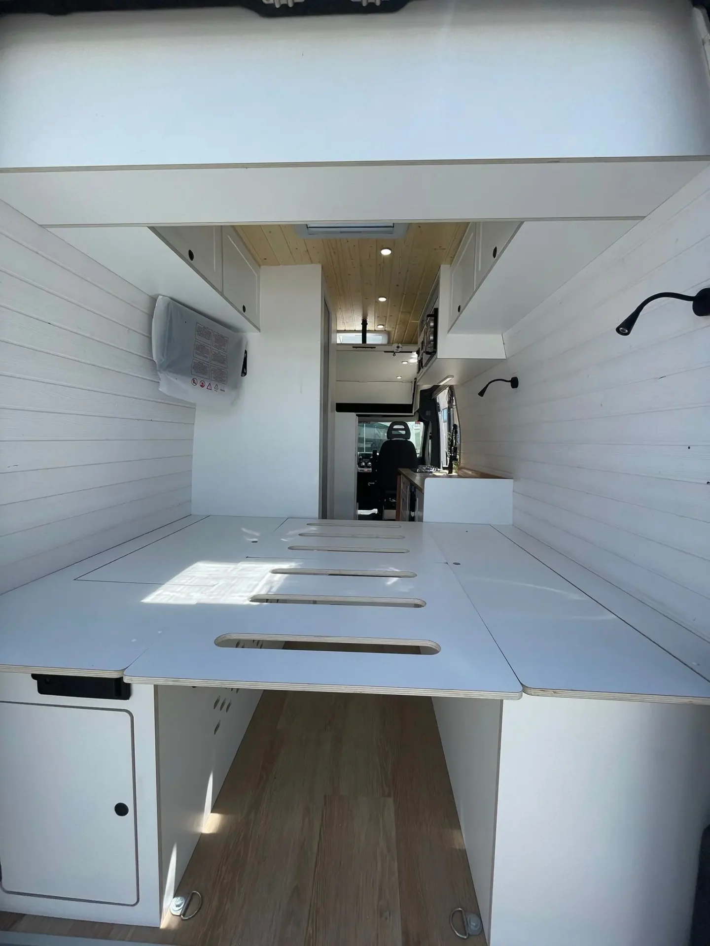 Interior de la furgoneta camperizada Ducato L4H3 con cama abatible, mostrando el corredor central con almacenamiento superior y espacio maximizado.