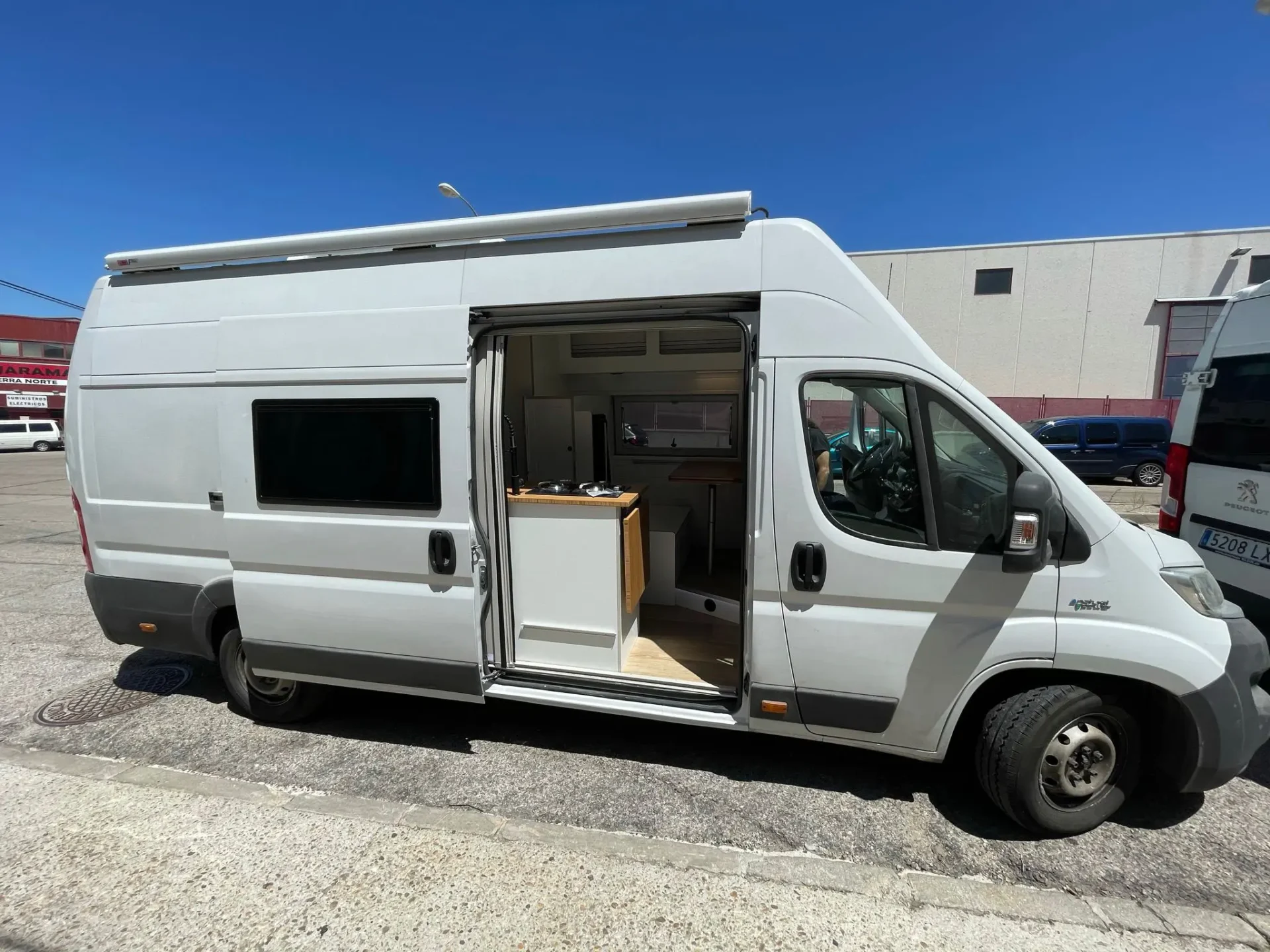 Furgoneta camper Ducato L4H3 aparcada, puertas abiertas revelando un diseño interior personalizado con cama abatible, cocina y zona de comedor, perfecta para viajes y aventuras.