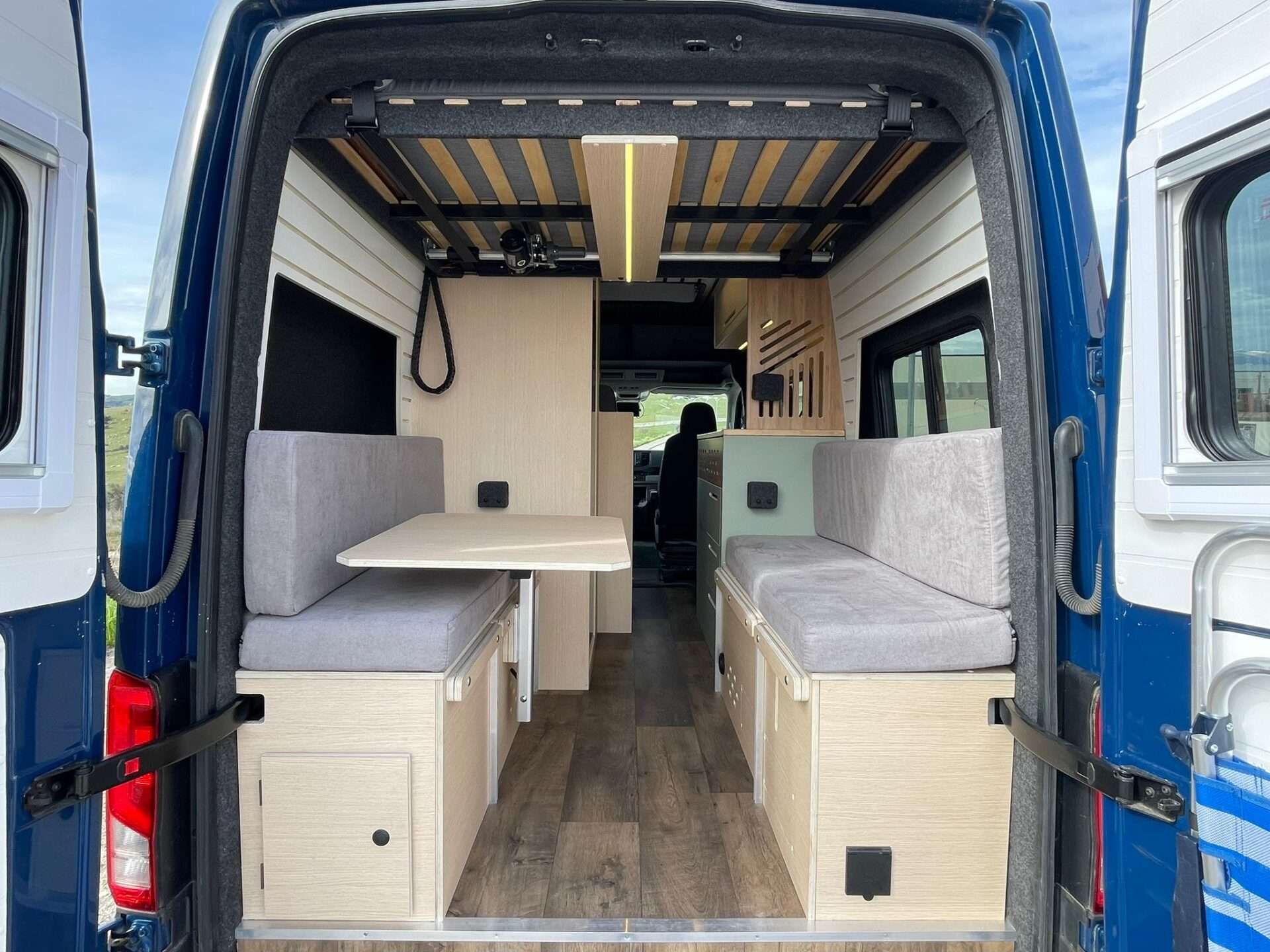 Camper Baovan con puertas traseras abiertas mostrando área de comedor con asientos y mesa, y almacenamiento inteligente.