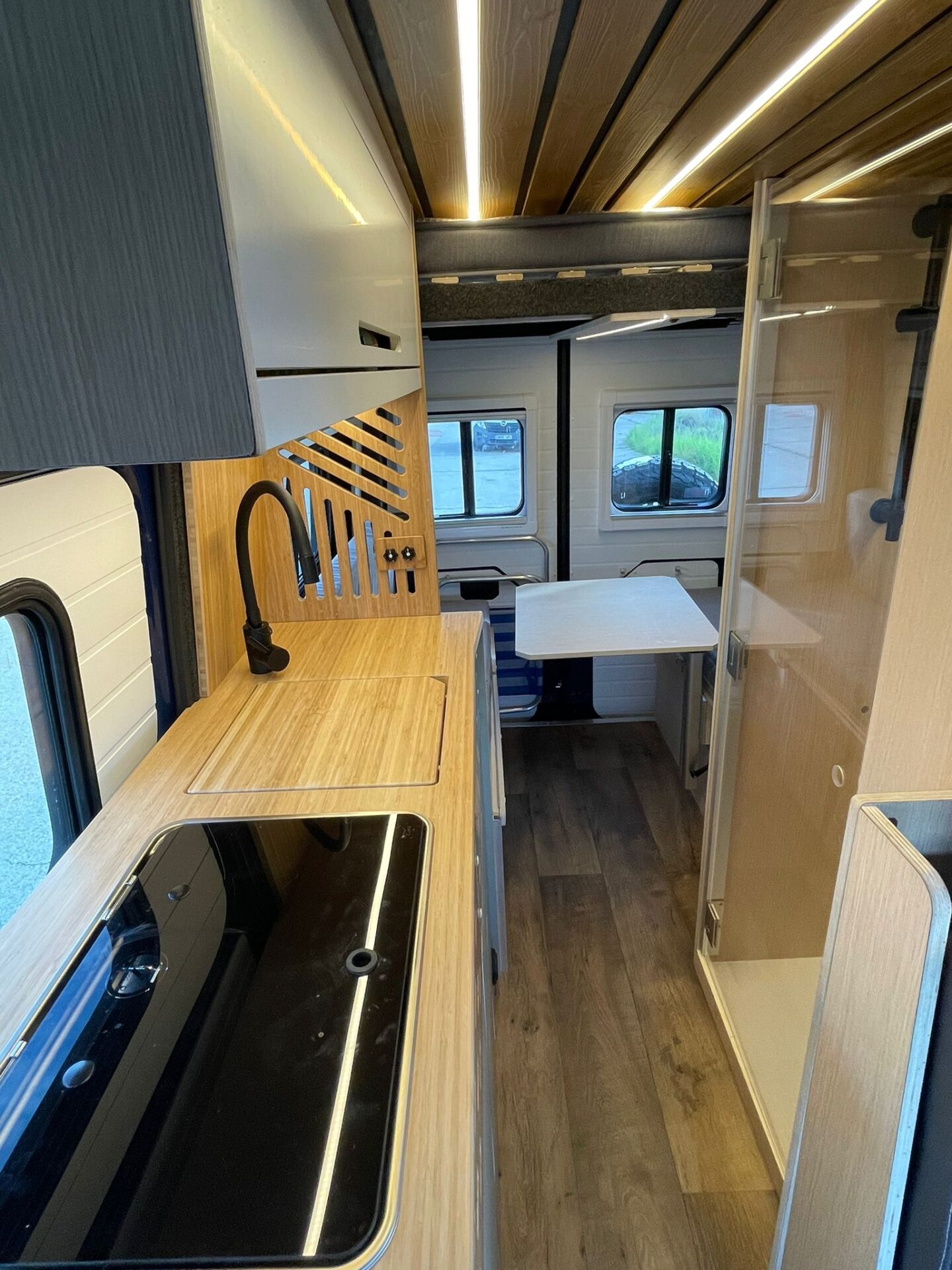 Cocina moderna en camper Baovan con encimera brillante, fregadero integrado, y mesa de comedor extensible adyacente a ventanas.