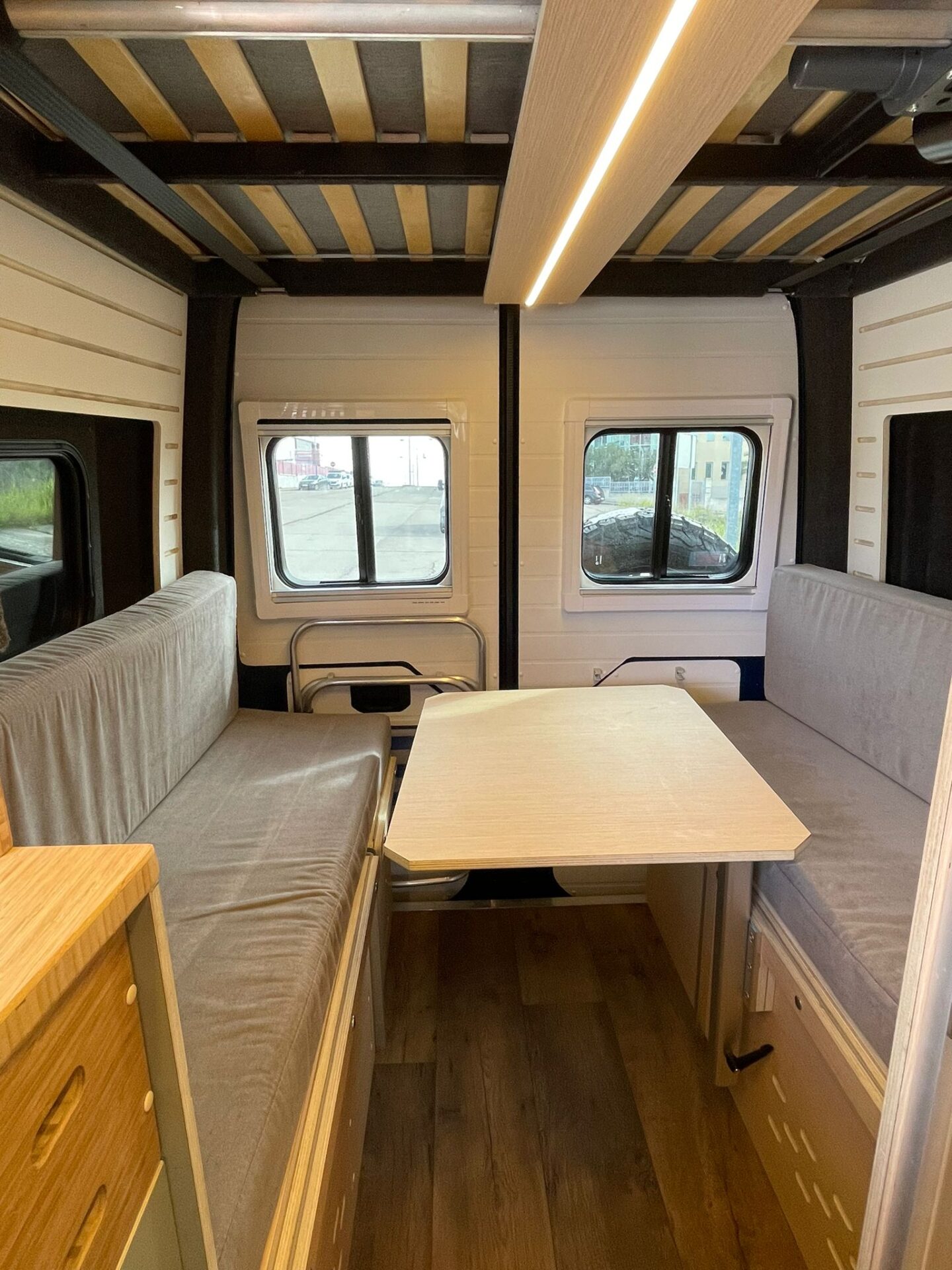 Área de comedor acogedora dentro de un camper Baovan con asientos grises a cada lado de una mesa central extensible y ventanas en ambos extremos.
